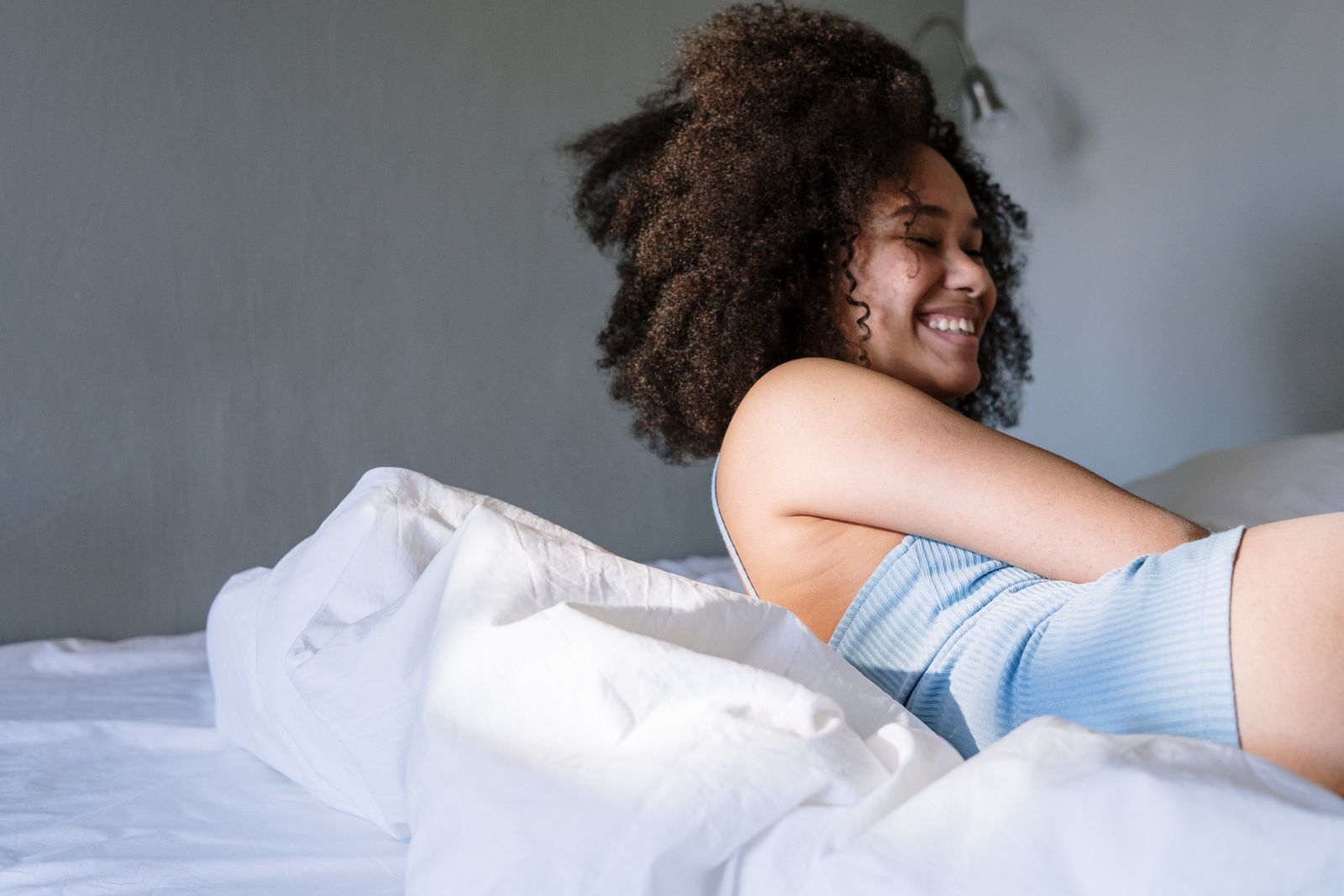 Woman with curly hair enjoys a calm morning in a cozy bedroom, smiling in the sunlight.