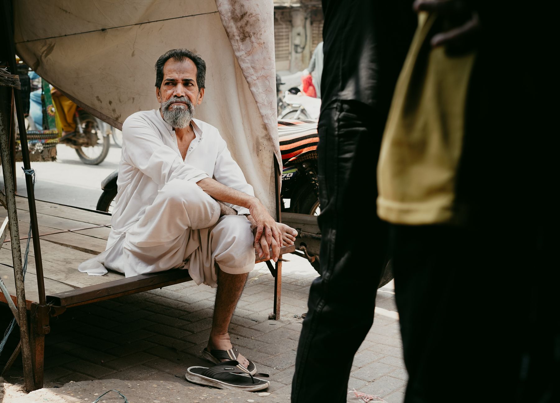 An elderly man in traditional attire sits thoughtfully on a street bench in Hyderabad, Pakistan.