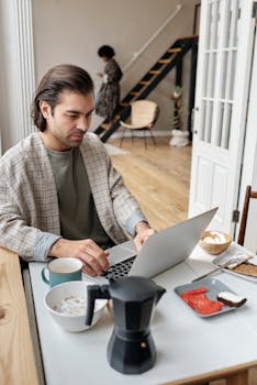 Man working remotely at breakfast table with coffee and laptop in cozy apartment setting.