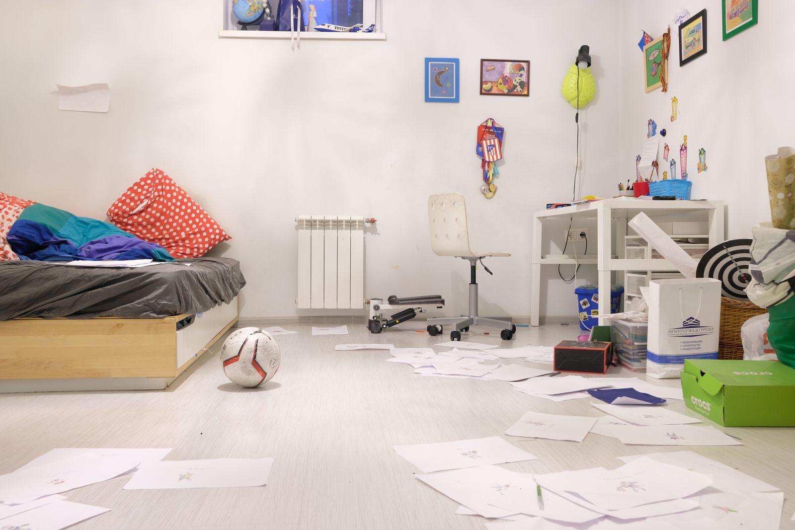 A child's bedroom with drawings scattered across the floor and a soccer ball by the bed, the room empty