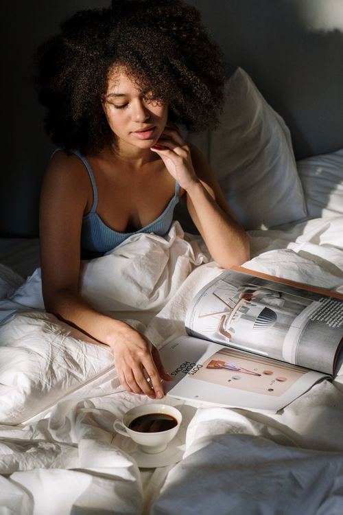 Free A woman with curly hair reads a magazine with coffee in a cozy bedroom setting. Stock Photo