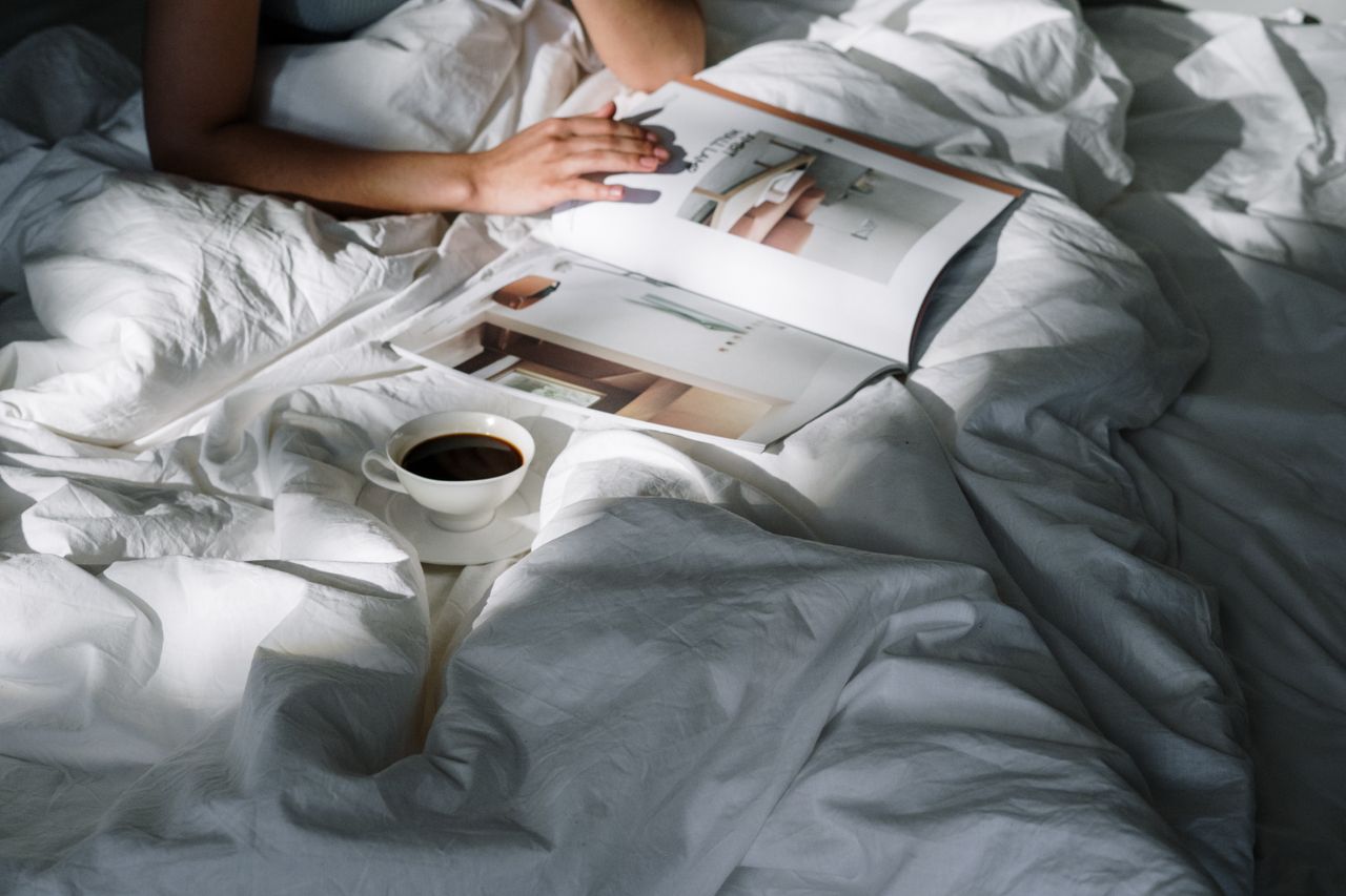 Hands holding a coffee cup in a linen-draped bed during slow morning light