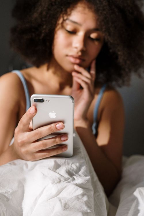 Free Young woman with curly hair using a smartphone in a cozy, sunlit bedroom. Stock Photo