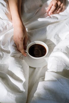 A person enjoys a cup of black coffee on white bed linens in a cozy bedroom.