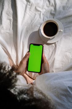 A woman relaxing in bed with a smartphone and coffee, enjoying a cozy morning at home.