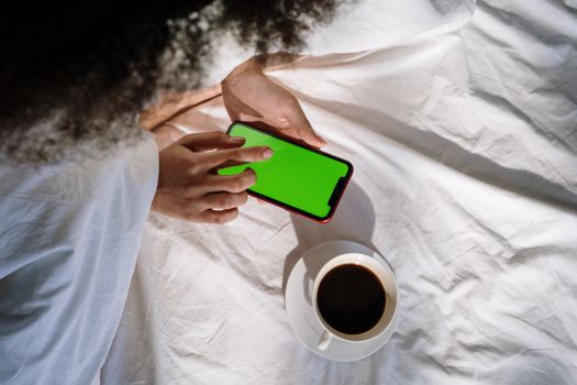A woman in bed using a smartphone with a cup of coffee nearby, creating a cozy morning atmosphere.