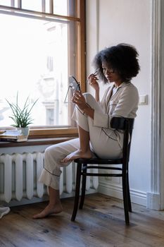 A woman sitting in a chair by the window applying makeup in a cozy home setting.