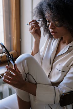 Woman applying skincare in pajamas near a window in natural light.