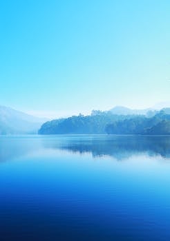A tranquil lake reflecting misty mountains under a bright blue sky, offering a sense of calm and serenity.