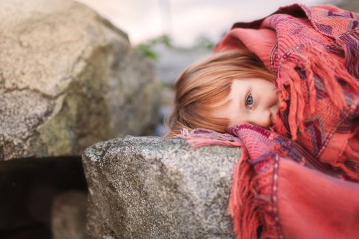 Portrait of a shy young girl wrapped in a red blanket, resting on rocks outdoors in Vancouver.