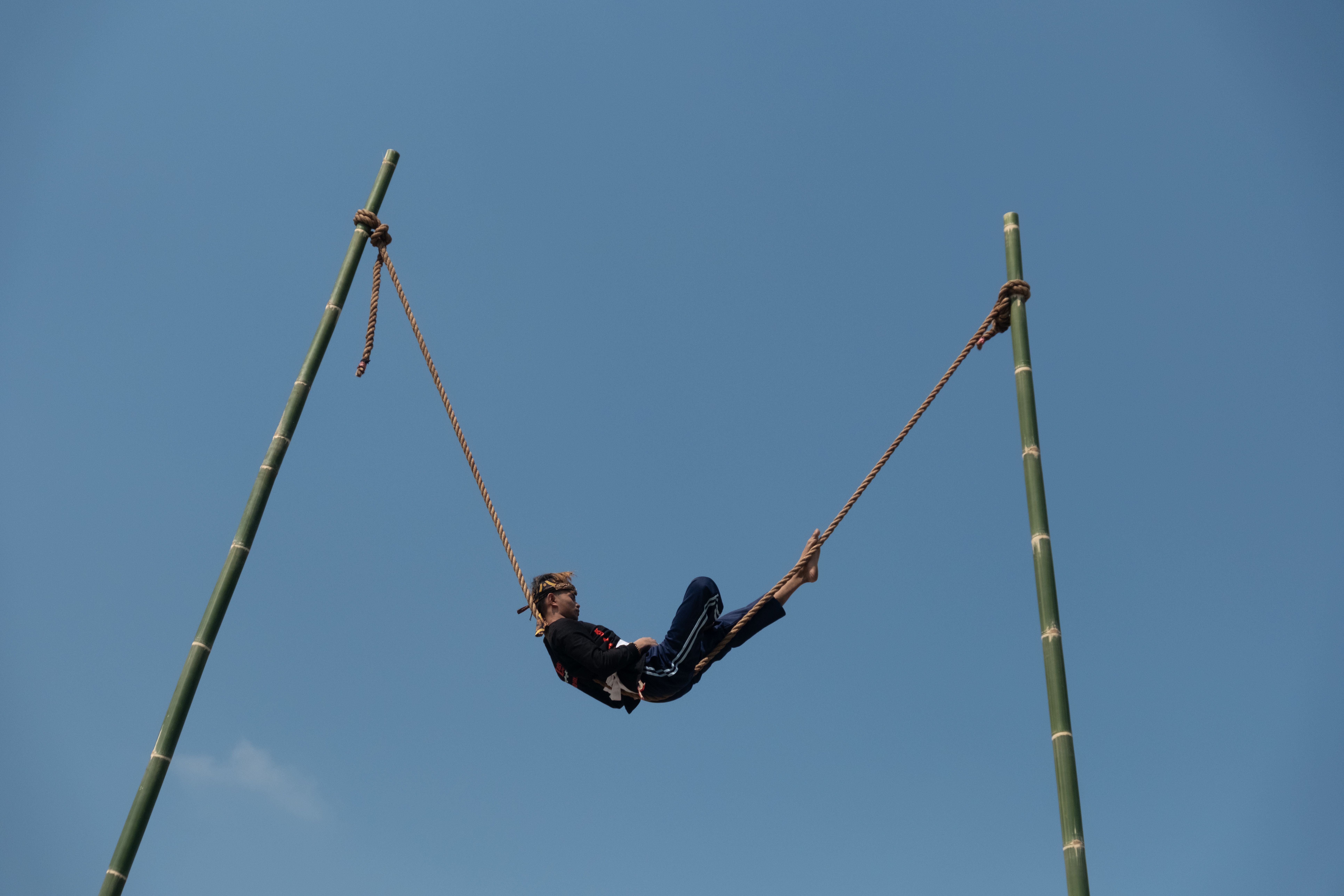 A man swings on a tall bamboo structure under a clear blue sky in West Java, Indonesia.