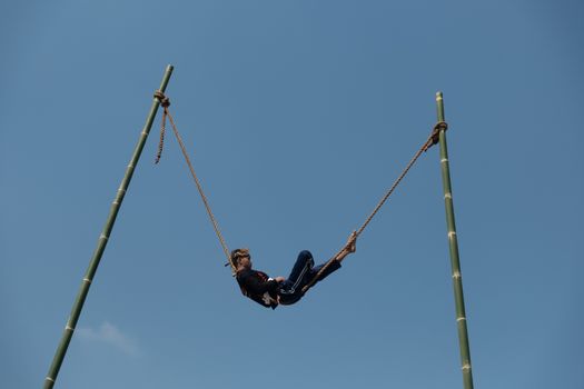 A man swings on a tall bamboo structure under a clear blue sky in West Java, Indonesia.
