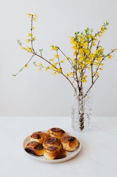 Plate of fresh cinnamon rolls beside a vase of yellow flowers, creating a delightful still life.