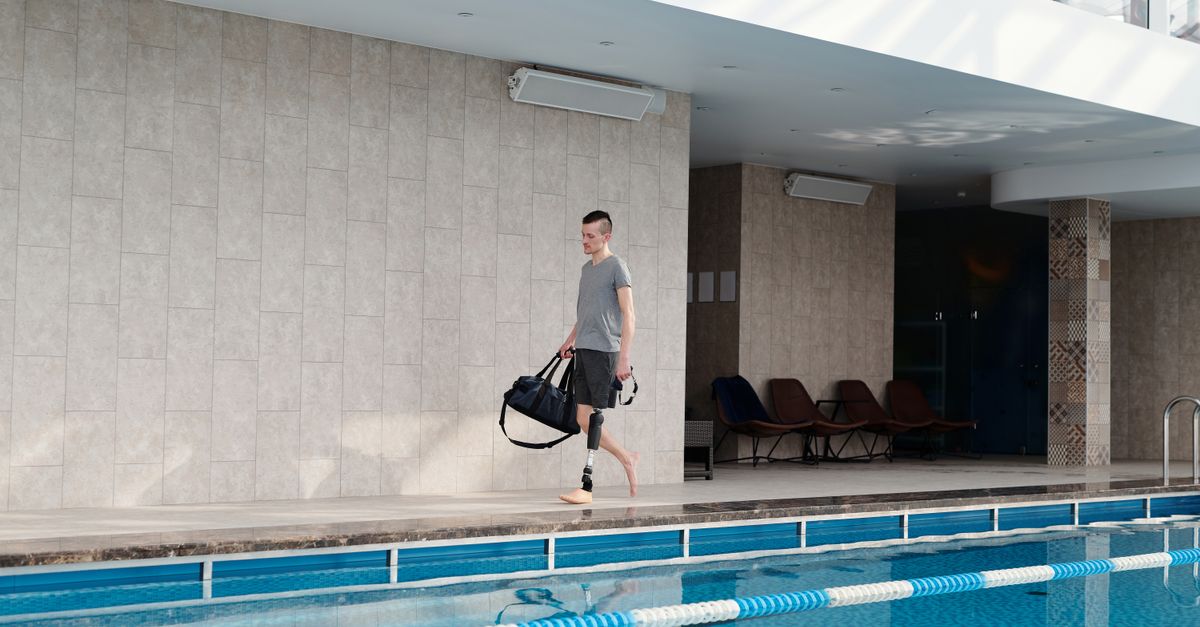 Man with prosthetic leg walking beside an indoor swimming pool carrying a gym bag.