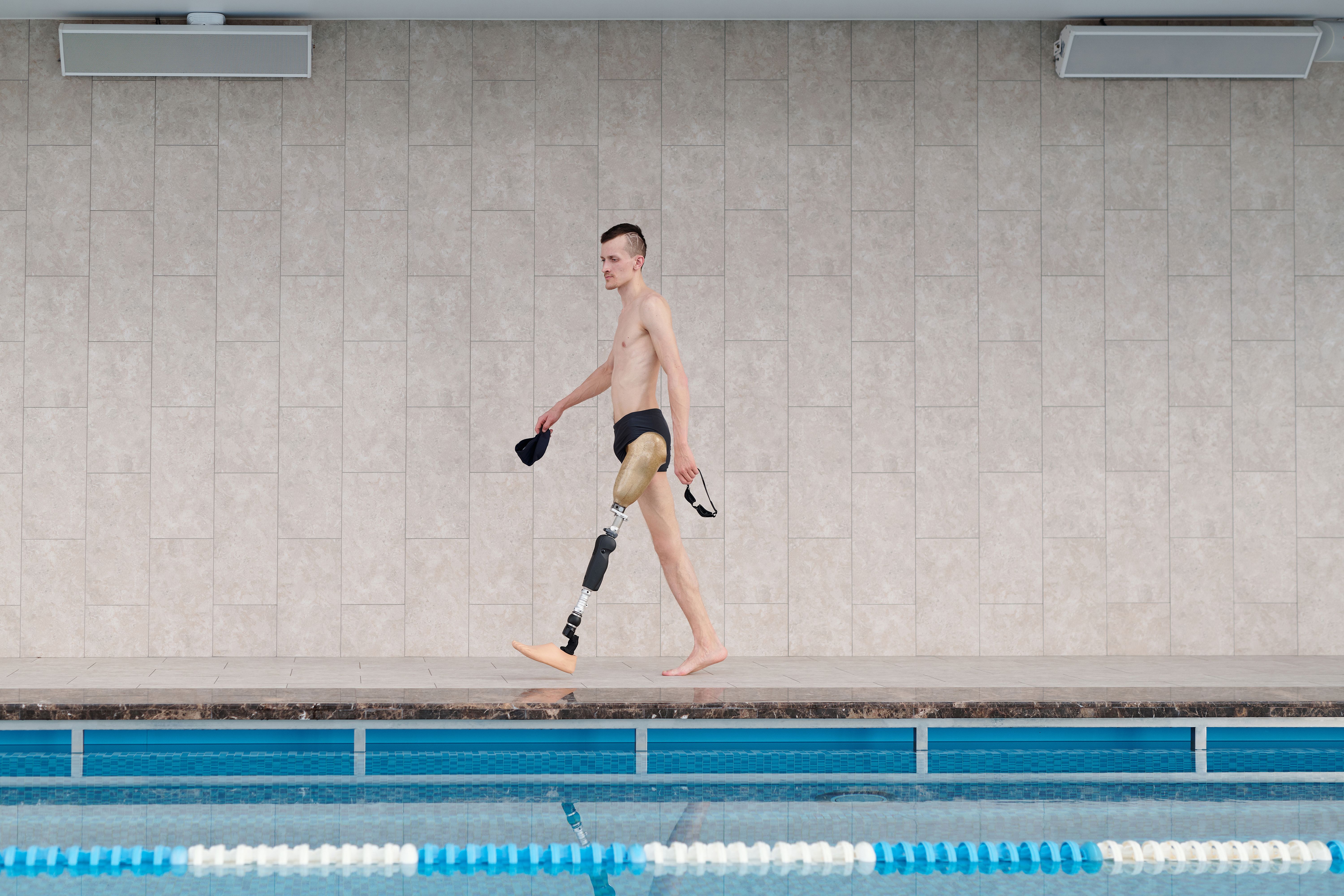 Amputee athlete with prosthetic leg walking by indoor swimming pool, holding goggles.