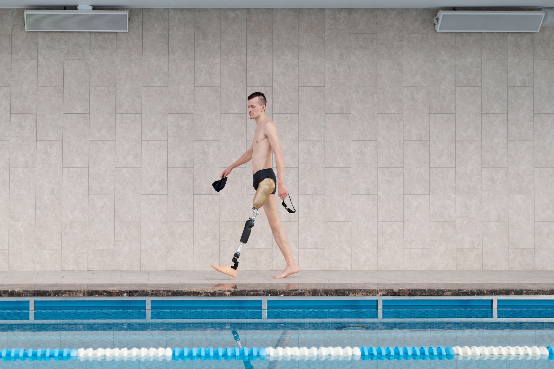 Amputee athlete with prosthetic leg walking by indoor swimming pool, holding goggles.