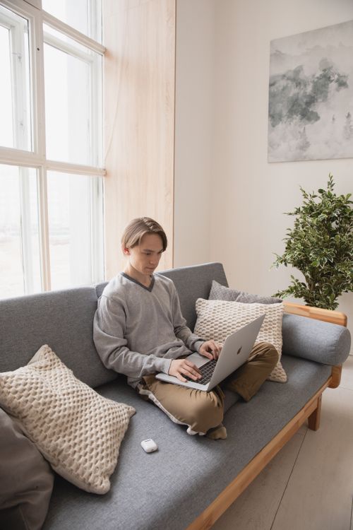 Free Young man using laptop while sitting on a cozy sofa in a bright living room. Stock Photo