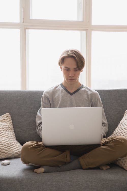 Young male remote employee browsing social media on portable computer while sitting with crossed legs on cozy sofa near window in afternoon in flat