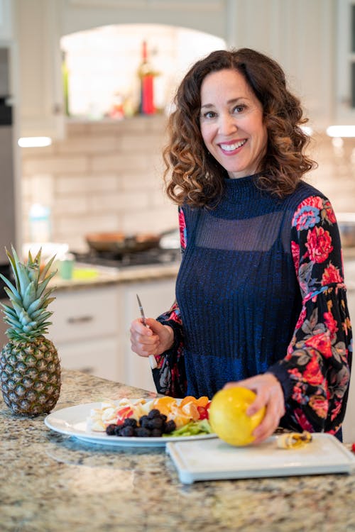 Free Cheerful woman standing near plate with tasty fruit slices with ripe mango on cutting board and pineapple aside during cooking process at home while looking at camera Stock Photo