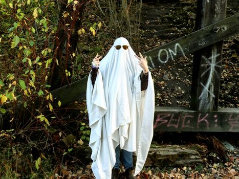Person in Halloween ghost costume with sunglasses, surrounded by fall foliage and graffiti.