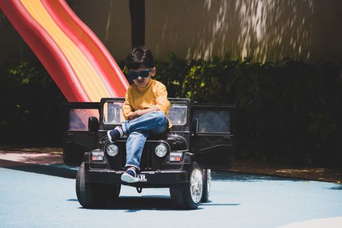 Free A young boy in sunglasses sits confidently on a toy car at an outdoor playground, enjoying a sunny day. Stock Photo