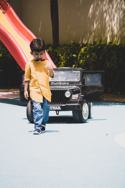 Free A young boy in sunglasses playing with a toy car outdoors near a playground slide. Stock Photo