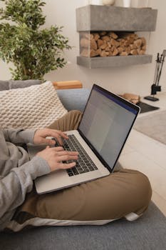 Person working on a laptop in a comfortable home setting with a modern fireplace.