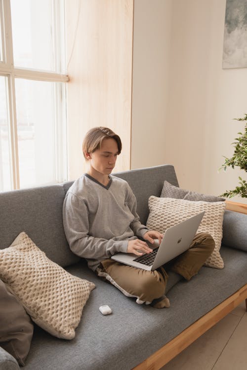 Young male freelancer typing on portable computer while sitting with crossed legs on cozy couch with cushions near window in apartment