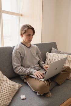 Cozy indoor scene of a young man working on a laptop from a couch, enjoying daylight in a comfortable setting.