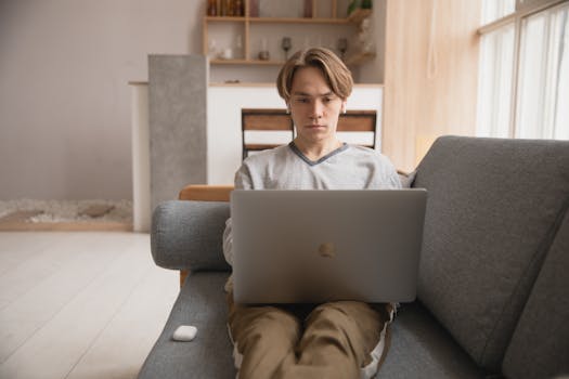A young man sitting on a sofa working remotely on a laptop in a cozy indoor setting.