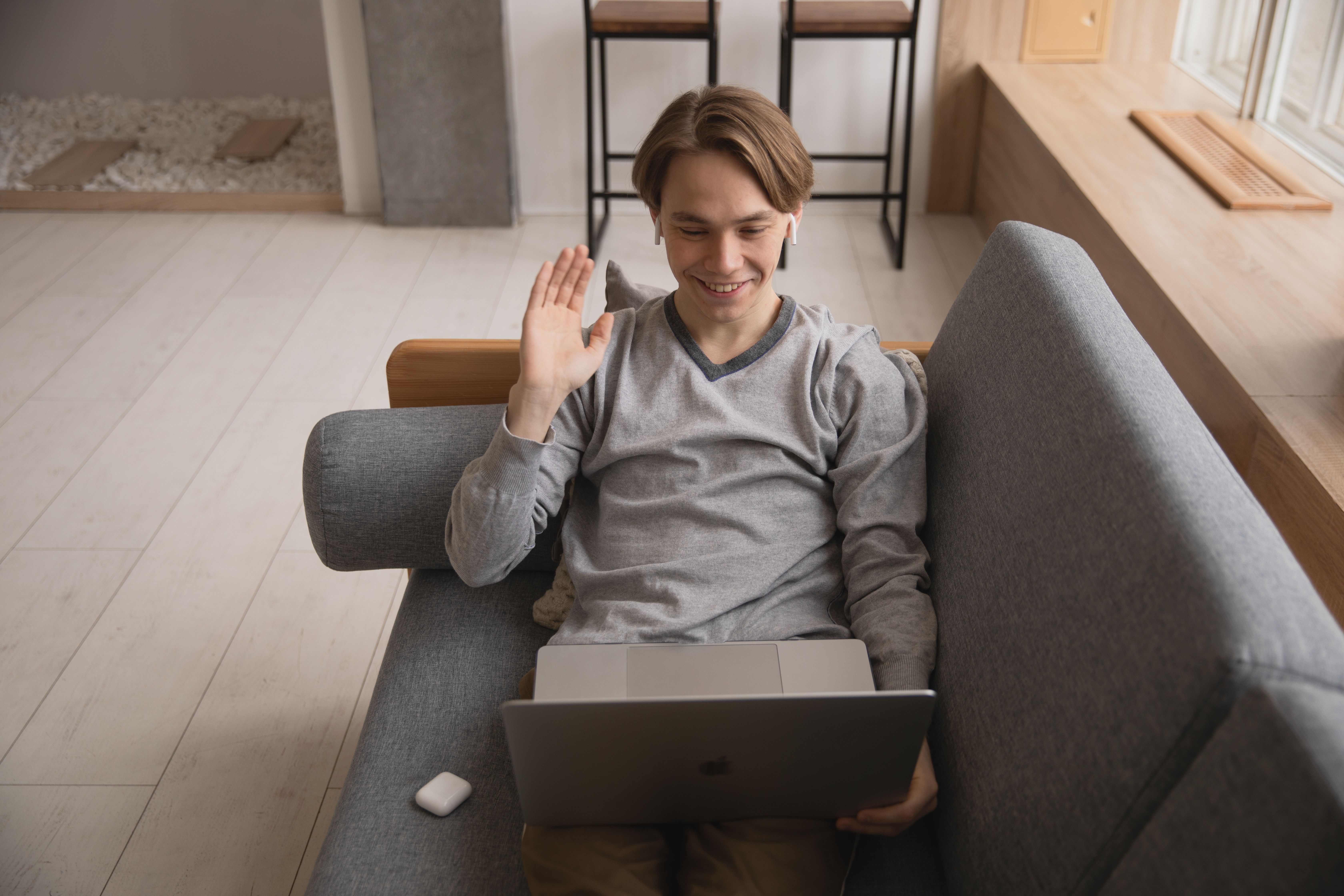 A smiling young man waves during a video call in front of his laptop at home