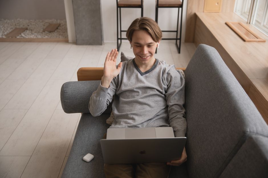 A smiling young man waves during a video call in front of his laptop at home