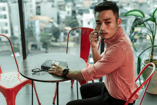 A fashionable man in a red checkered shirt talks on his phone in a modern cafe setting.