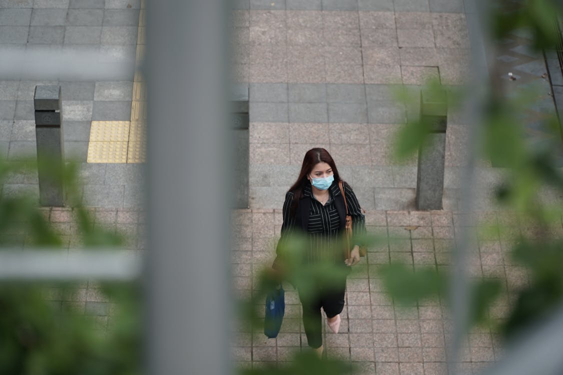 Free From above of anonymous woman in formal wear with shoulder bag in sterile mask strolling on tiled pavement on city street and looking away during quarantine period Stock Photo