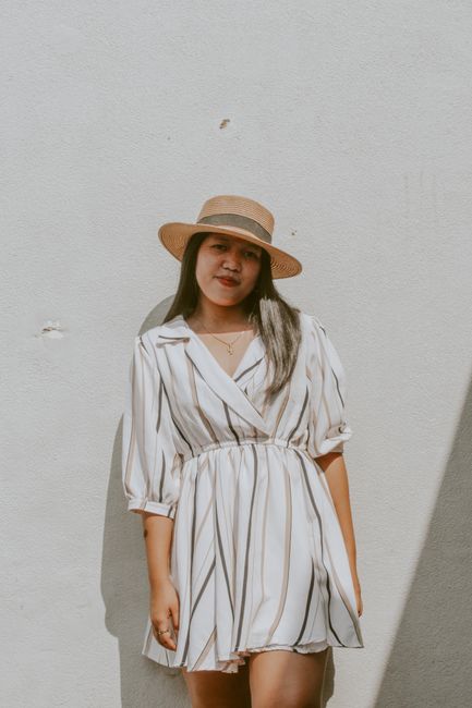 A fashionable woman wearing a striped dress and straw hat poses against a textured wall in sunlight.