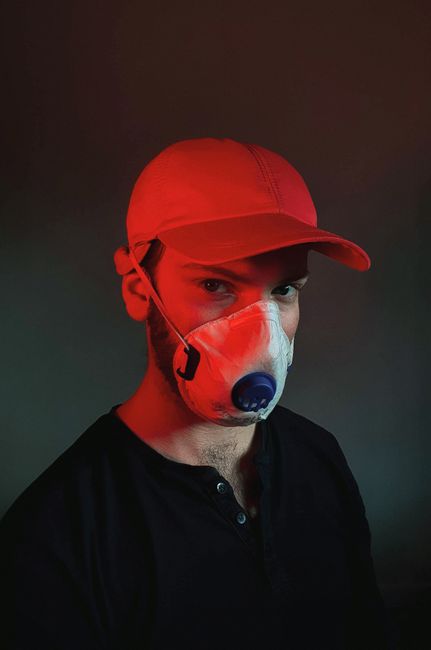 Portrait of a man wearing a mask and red baseball cap, illuminated by dramatic lighting.