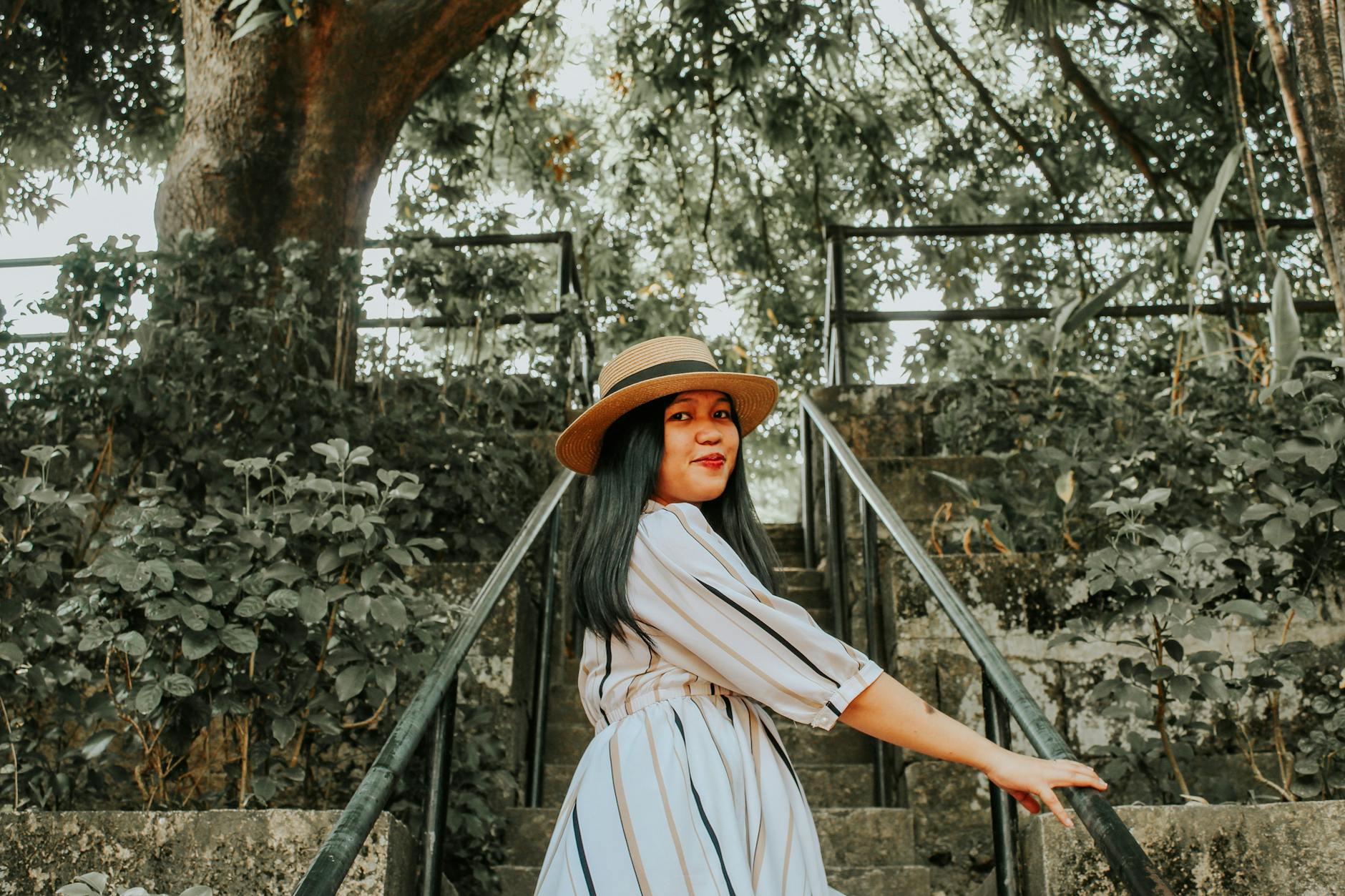 A fashionable woman poses with a straw hat on outdoor steps surrounded by greenery.