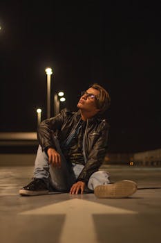 Stylish man in a leather jacket sitting on a road at night, looking contemplative.