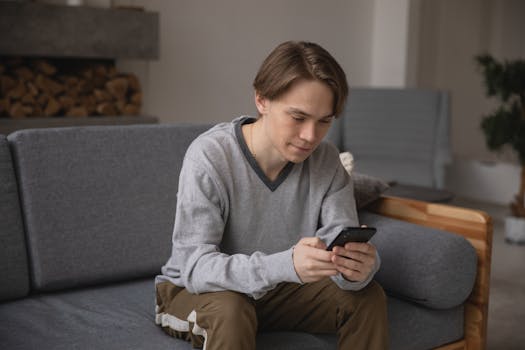 A young man comfortably sitting on a gray couch using his smartphone indoors.
