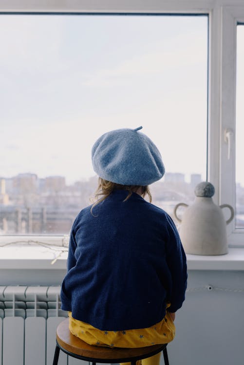 Back view of anonymous kid wearing blue beret sitting on stool in modern apartment and looking on blurred city exterior near windowsill with creative composition of ceramic vase and gray stone and willow twig in spring day