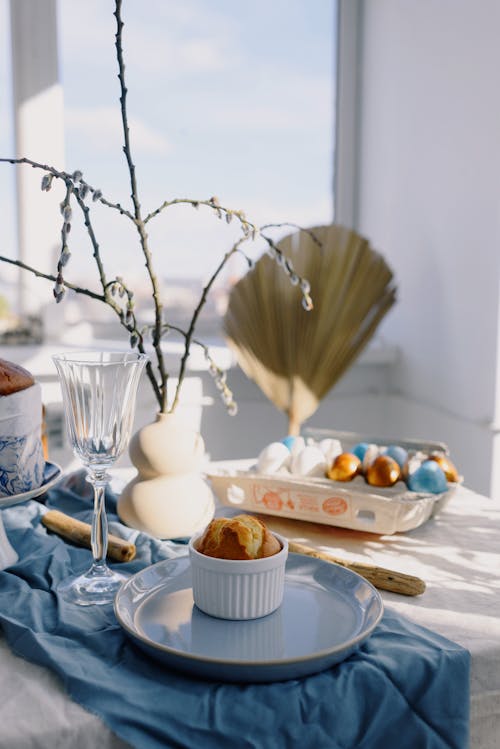 Festive table setting with colored eggs and Easter cake placed on blue napkin with spring willow twig in vase against window with bright blue sky
