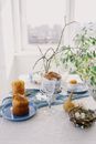 Festive table setting with colored eggs and Easter cakes on blue napkin with willow twig in vase and decorative nest with eggs placed on white tablecloth during sunny spring day