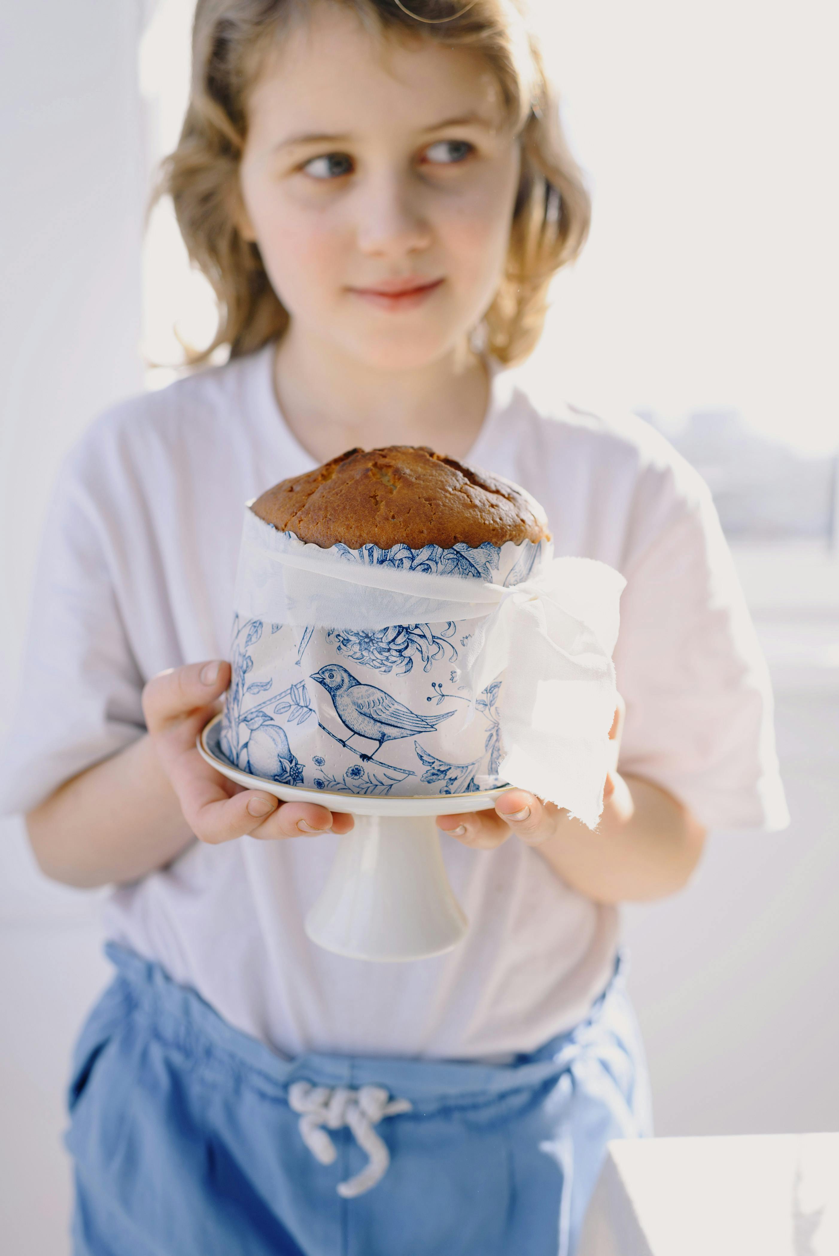Cheerful little girl in casual clothes holding plate with delicious Easter cake in white paper mold with blue bird picture in light room