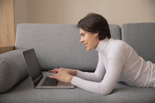Woman working remotely on a laptop at home, lying on a comfortable sofa.