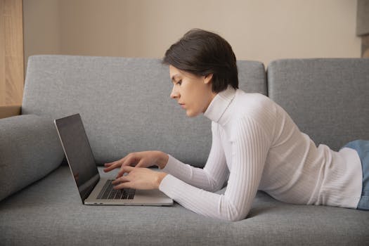 Young adult woman using laptop on a gray sofa indoors, engaged in remote work or leisure activities.