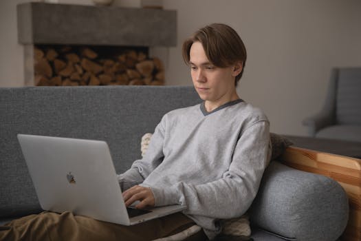 A young man in a comfortable home setting, using a laptop for remote work on a couch.