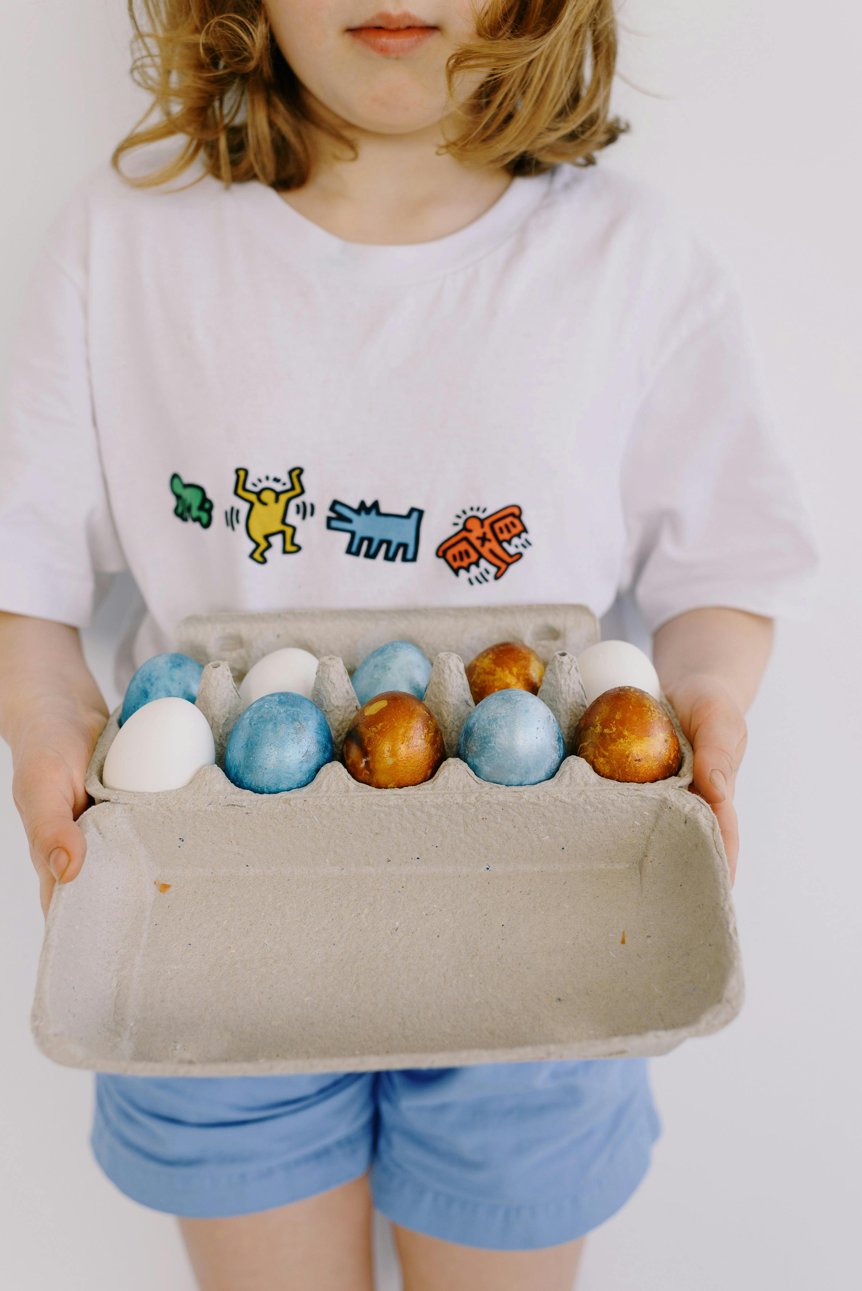 Little girl holding a carton of blue and brown Easter eggs wearing a graphic print shirt.
