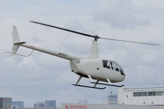 White Robinson R44 helicopter flying over city rooftops with cloudy sky.