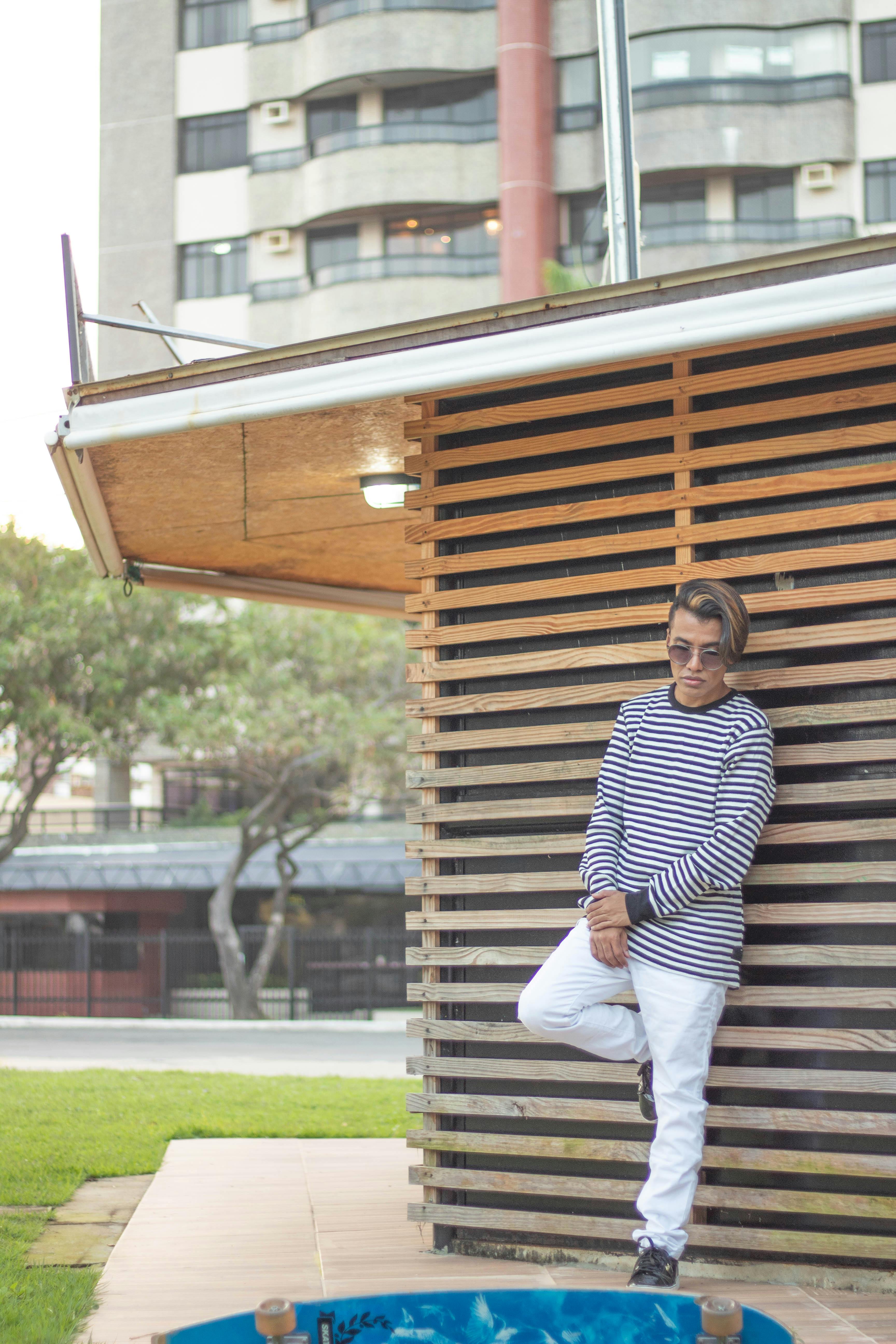 Slim stylish unrecognizable ethnic male with modern haircut in sunglasses standing leaned on one leg near wooden fence and green meadow with trees behind building facade in town in daylight