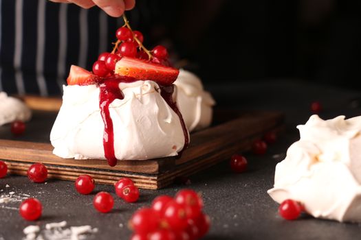 Crop anonymous chef decorating appetizing sweet meringue cake with juicy strawberry slices and sprig of colorful red currants on cutting board on table in cafe